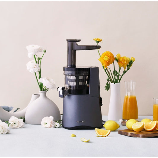 Hurom HA-2600 Classic Series slow juicer displayed alongside fruits and flowers, emphasizing its color and design.
